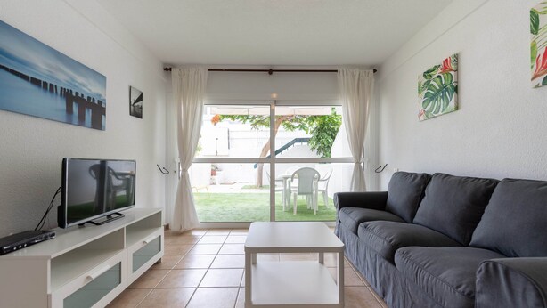 A living room with a TV, white coffee table, grey sofa, and a sliding glass door leading to a patio with outdoor furniture. - Apartment in San Agustin Near the Beach (San Bartolomé de Tirajana)