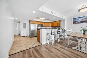 A modern kitchen with wooden cabinets, a stainless steel refrigerator, and a microwave. There is a dining area with white chairs and a wooden table.
