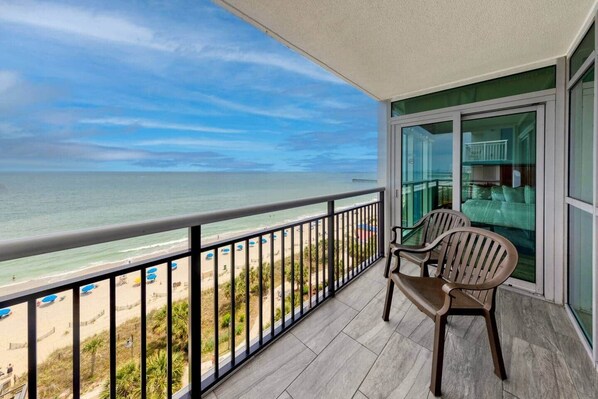 A balcony with a view of the beach and ocean, a chair, and a glass door leading to a room.