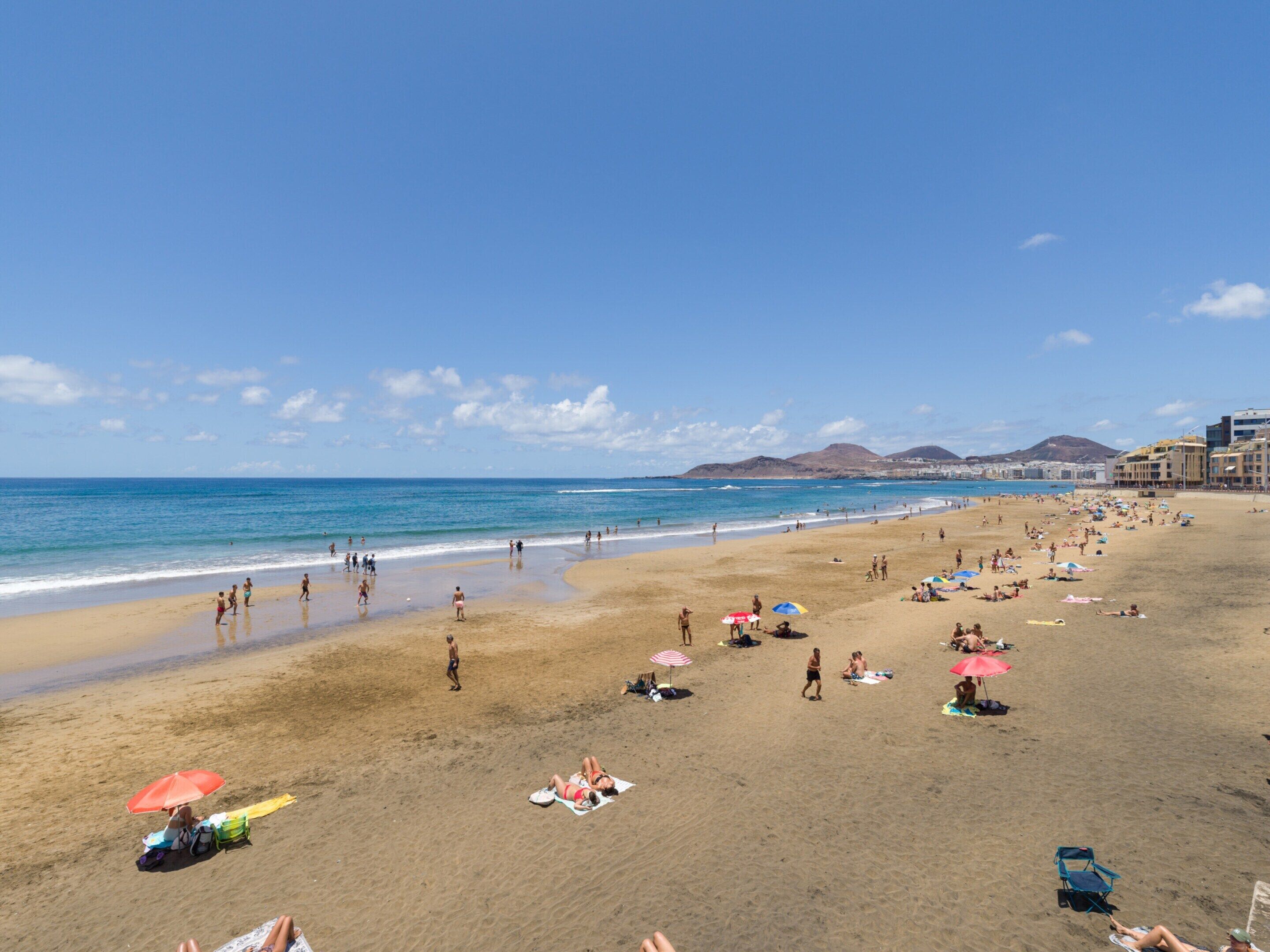 A beach with people sunbathing and swimming, a clear sky, and buildings in the background.