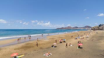 A beach with people sunbathing and swimming, a clear sky, and buildings in the background.