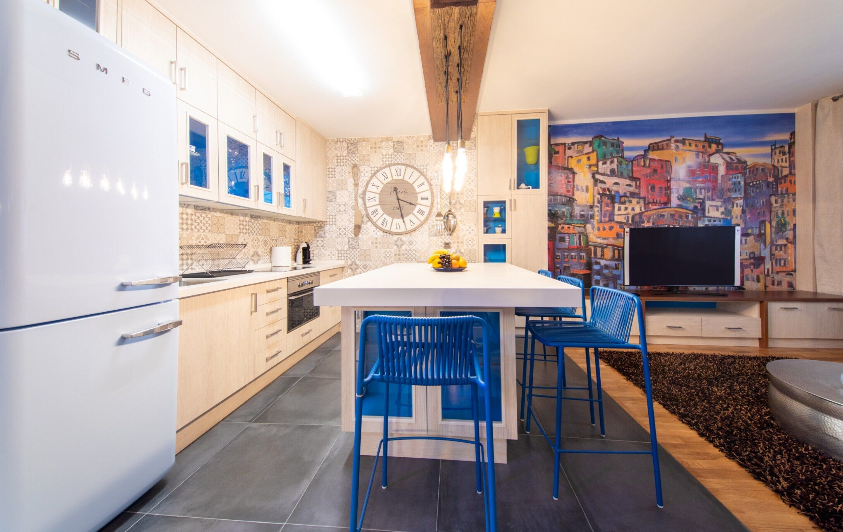 A modern kitchen with white cabinets, a central island with blue chairs, a large clock, and a colorful cityscape mural.