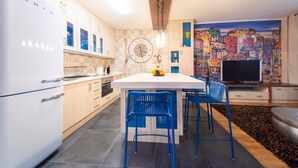 A modern kitchen with white cabinets, a central island with blue chairs, a large clock, and a colorful cityscape mural. - Apartment in Las Palmas Near Las Canteras Beach (Las Palmas de Gran Canaria)