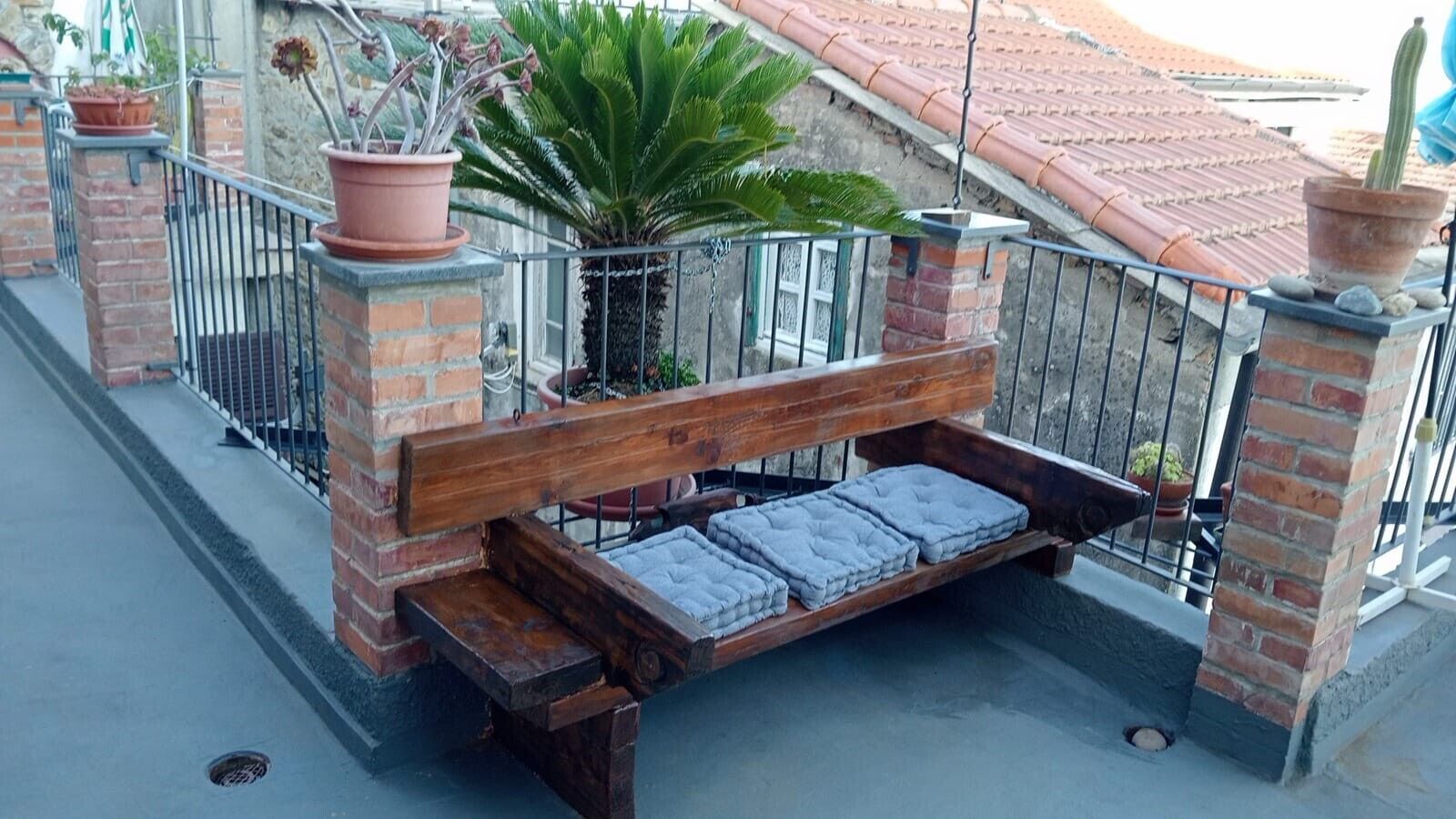 A wooden bench with cushions, surrounded by brick pillars and metal railings, with potted plants and a building in the background.