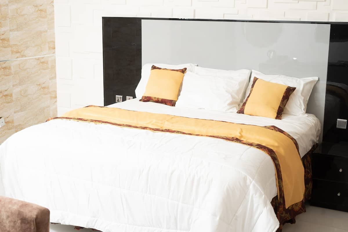 A neatly made bed with white and yellow bedding, a black headboard, and a white brick wall.