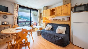 A compact kitchen with wooden cabinets, a microwave, and a refrigerator. There is a dining area with a wooden table and chairs, and a bed with pillows.
