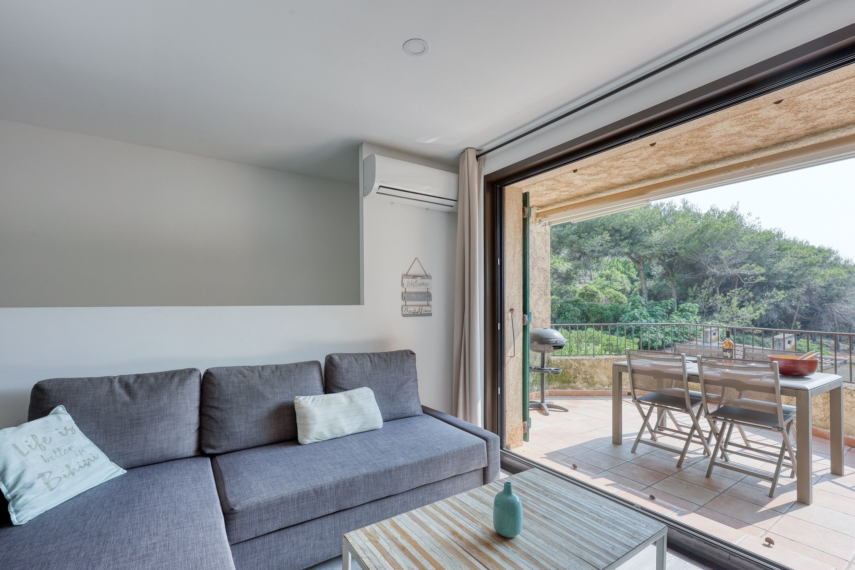 Un salon moderne avec un canapé gris, une table basse en verre et un balcon aménagé avec du mobilier d’extérieur.