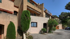A two-story building with a stone facade, a balcony, and a small garden area with a tree and shrubs.