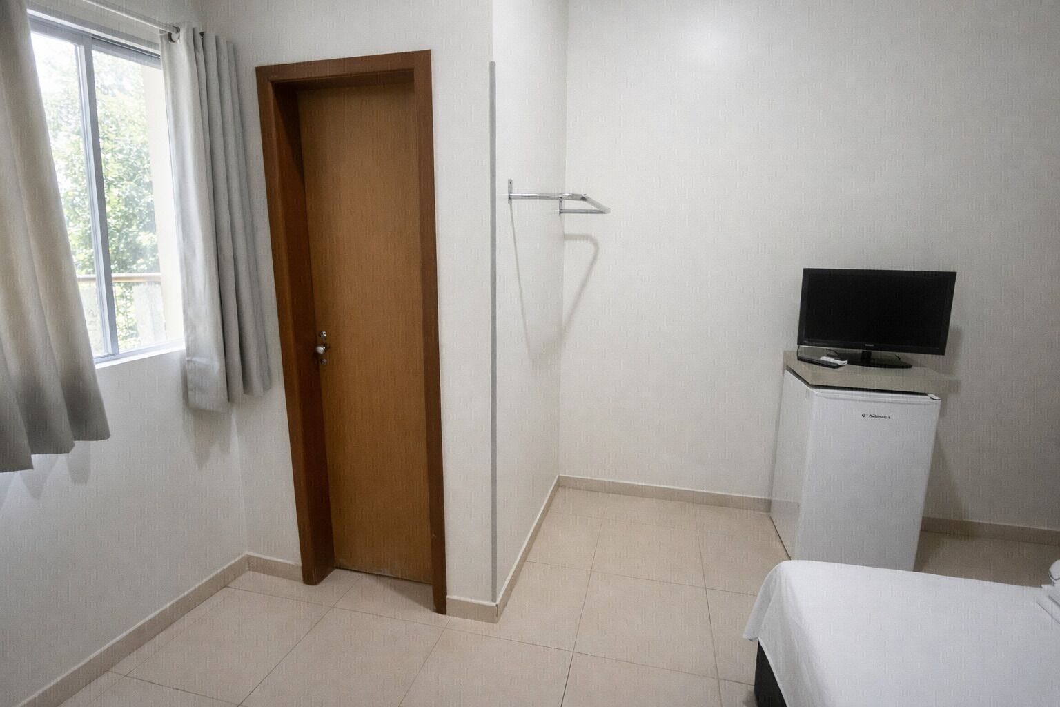 Une pièce avec une porte en bois, un réfrigérateur blanc, un téléviseur posé sur le dessus, et un lit recouvert d’une couverture blanche.