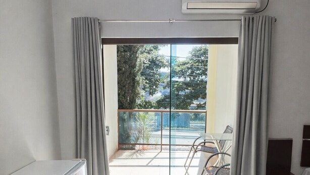 A modern living room with a sliding glass door leading to a balcony, a white air conditioning unit, a small white refrigerator, and a brown sofa. - Hotel Royal - Rubiataba (Rubiataba)