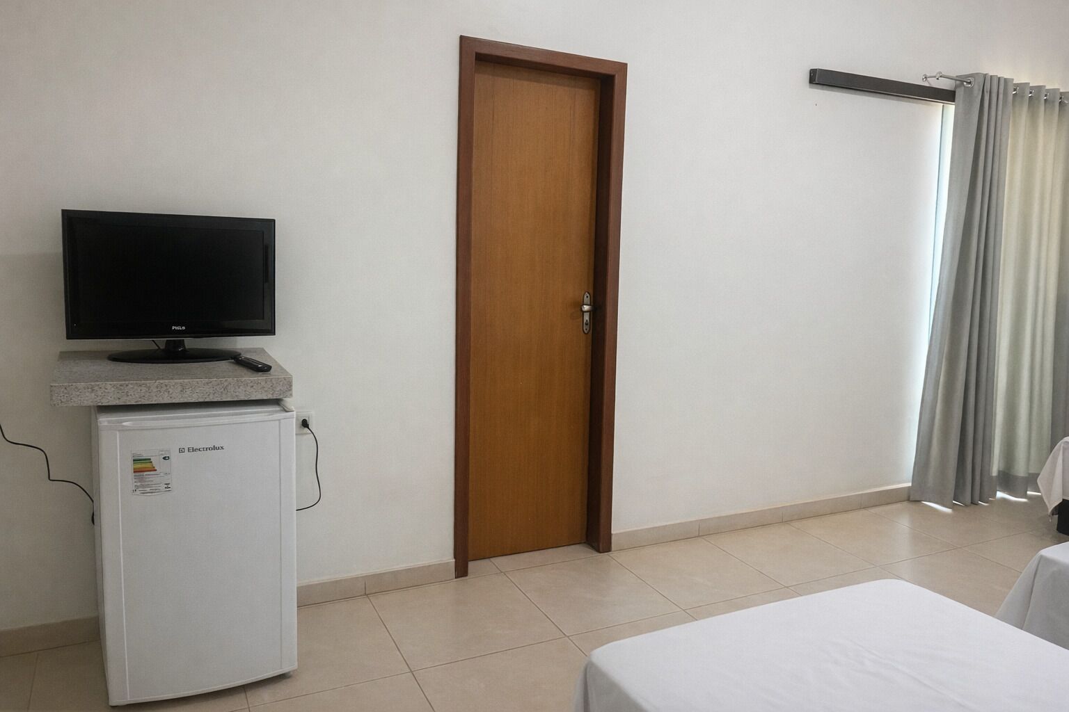 Une chambre d’hôtel avec un téléviseur posé sur une petite commode, un réfrigérateur, une porte en bois et un lit recouvert d’une couverture blanche.