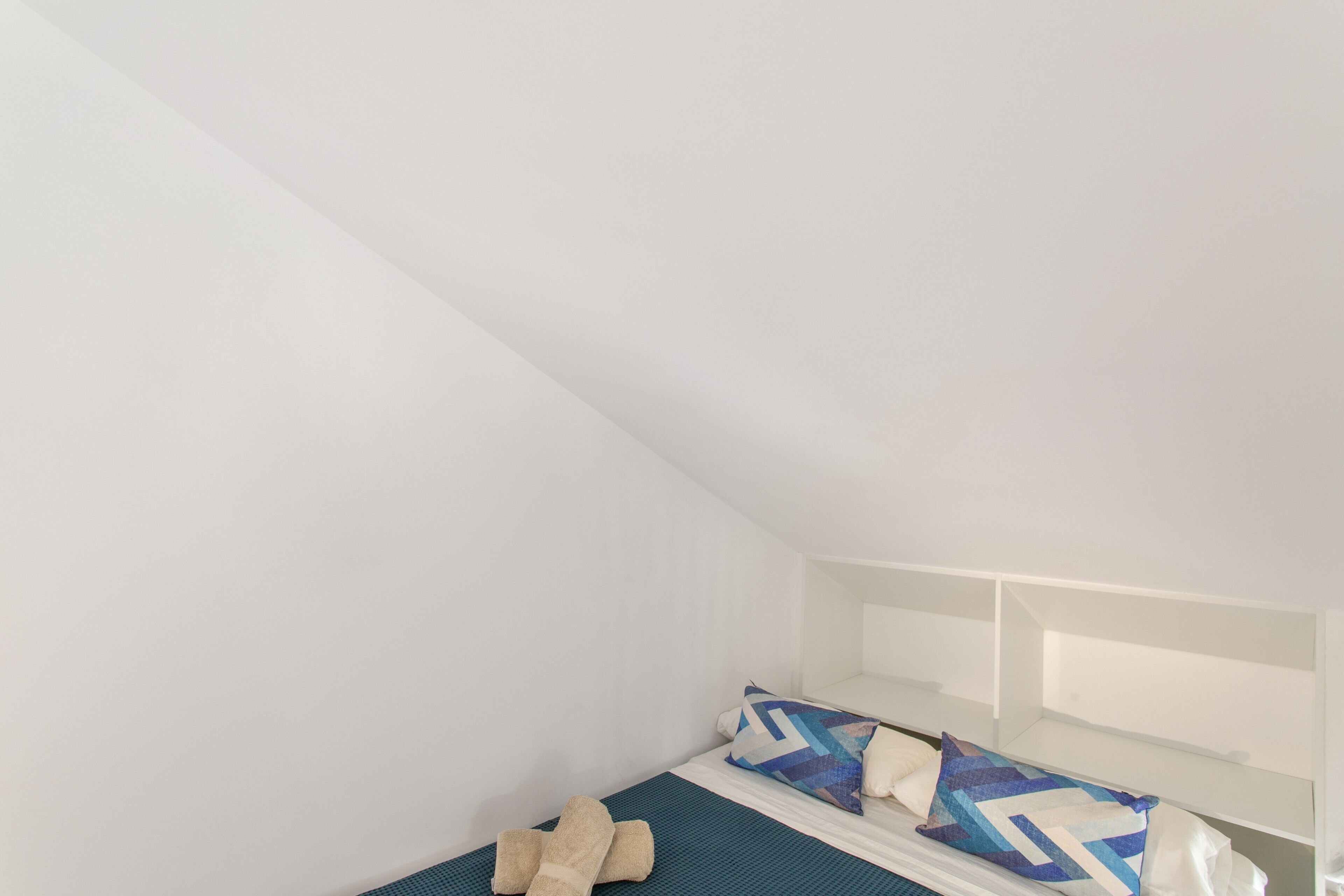Une petite chambre blanche avec un lit simple, un plafond en pente, un matelas bleu et deux oreillers à motifs géométriques.