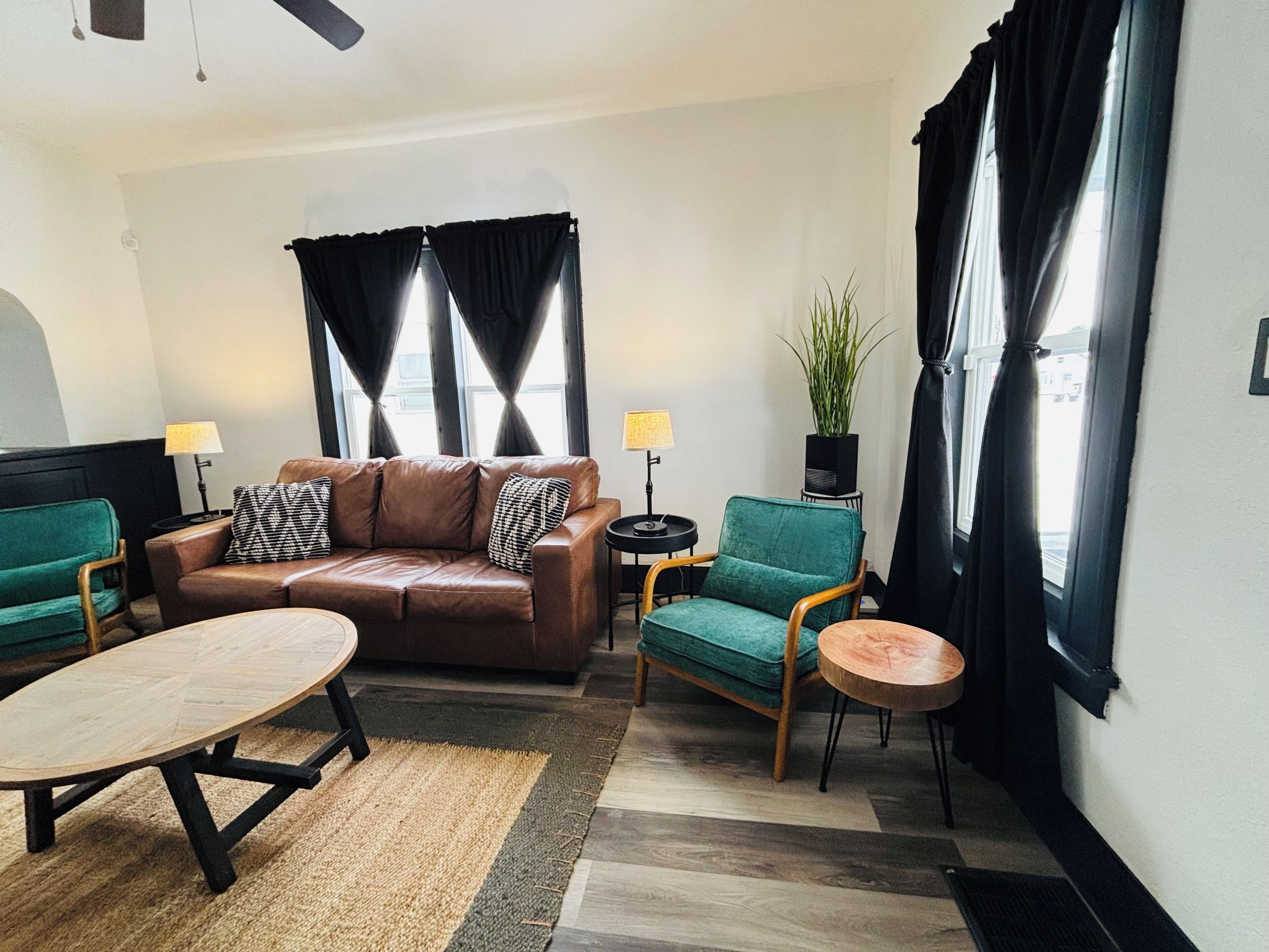 Un salon avec un canapé en cuir marron, deux fauteuils verts, une table basse ronde et une petite table d’appoint avec une lampe.
