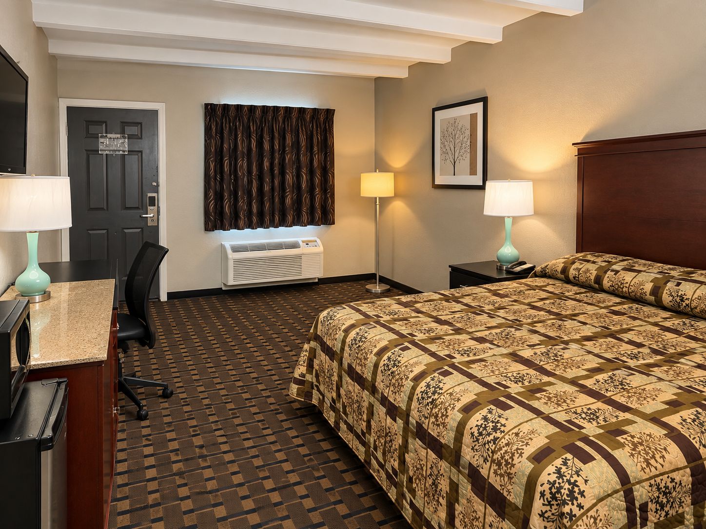Una habitación de hotel con cama, escritorio, silla, dos lámparas de noche, unidad de aire acondicionado y una puerta.