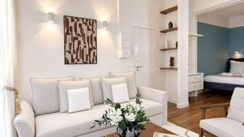 A modern living room with a sofa, a coffee table with a vase and book, and a bedroom with a bed and shelving.