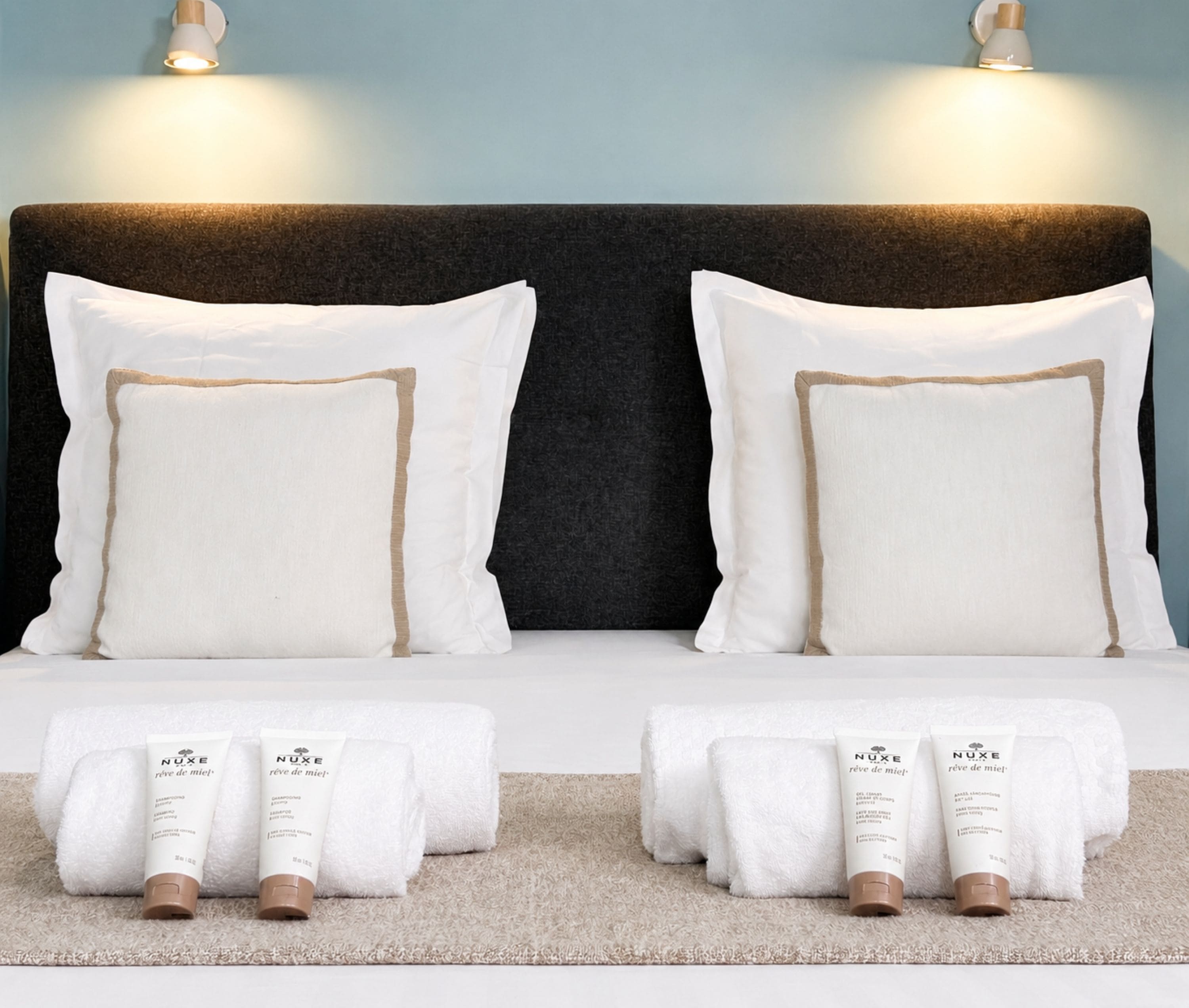 A neatly made bed with white pillows and two Nuxe shampoo bottles on a beige rug.