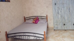 Una cama individual con estructura metálica y cabecera de madera en una habitación con paredes beige y suelo de baldosas.