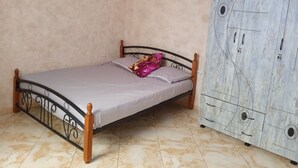 Una cama individual con estructura metálica y patas de madera, un colchón sencillo y una almohada decorativa rosa. A la derecha, un armario grande con diseño geométrico.