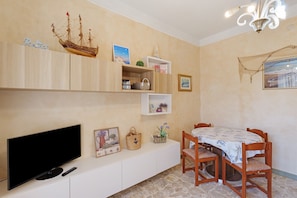 A room with a flat-screen TV, a dining table with chairs, a chandelier, and shelves with various items.