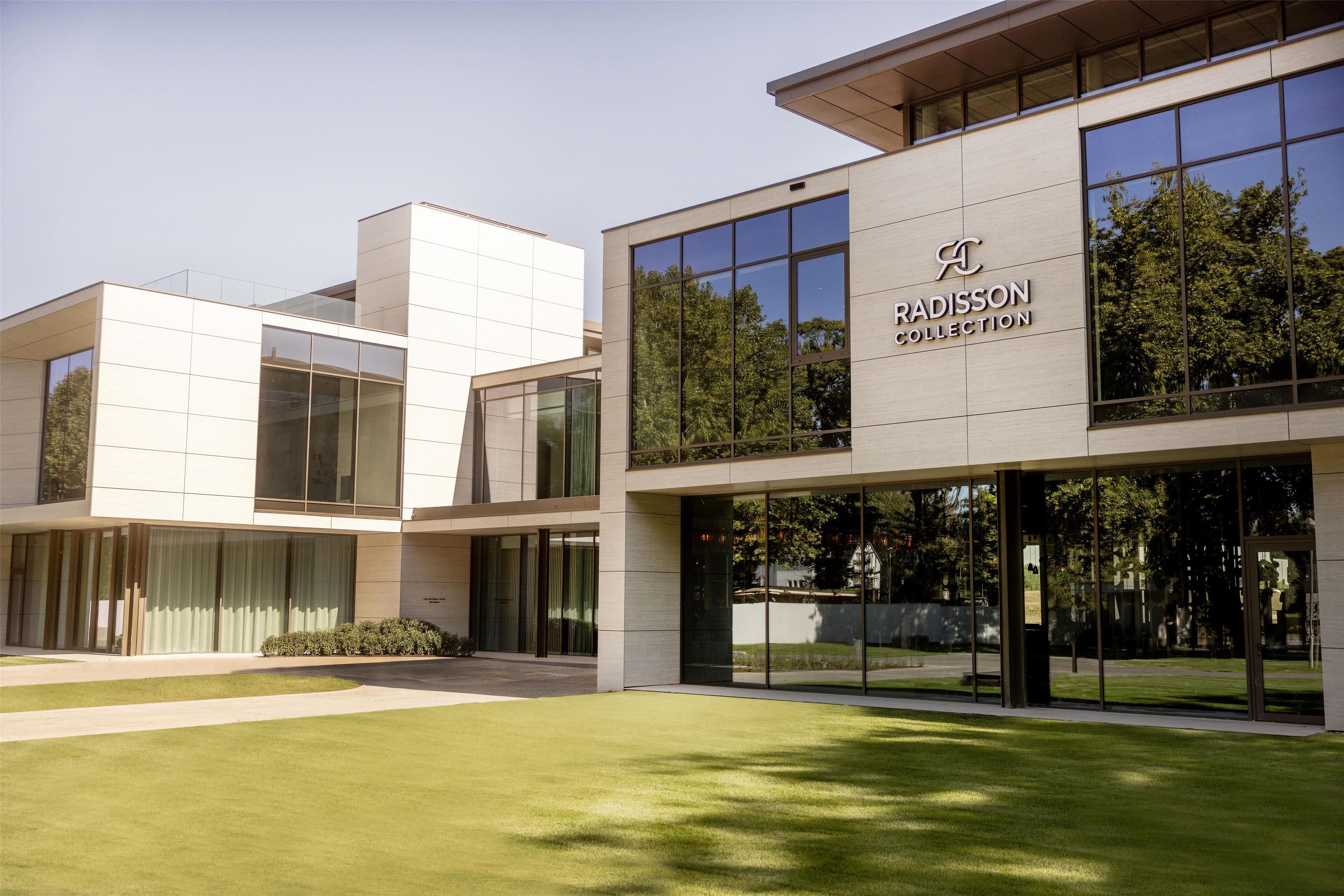 A modern building with a glass facade, a sign reading 'RADISSON COLLECTION', and a well-maintained lawn in front.