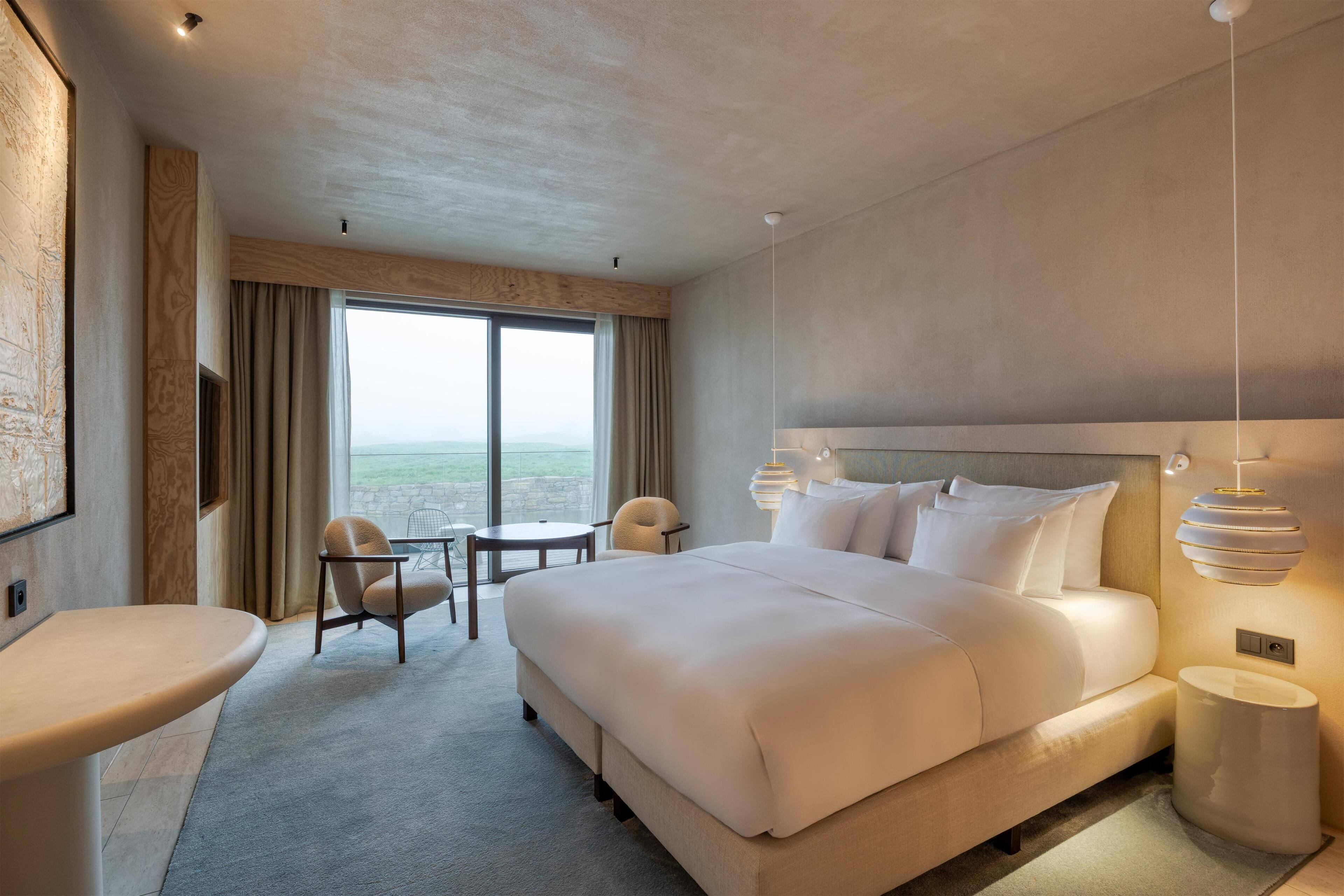 A modern hotel room with a large bed, a desk, a chair, and a view of the outdoors.