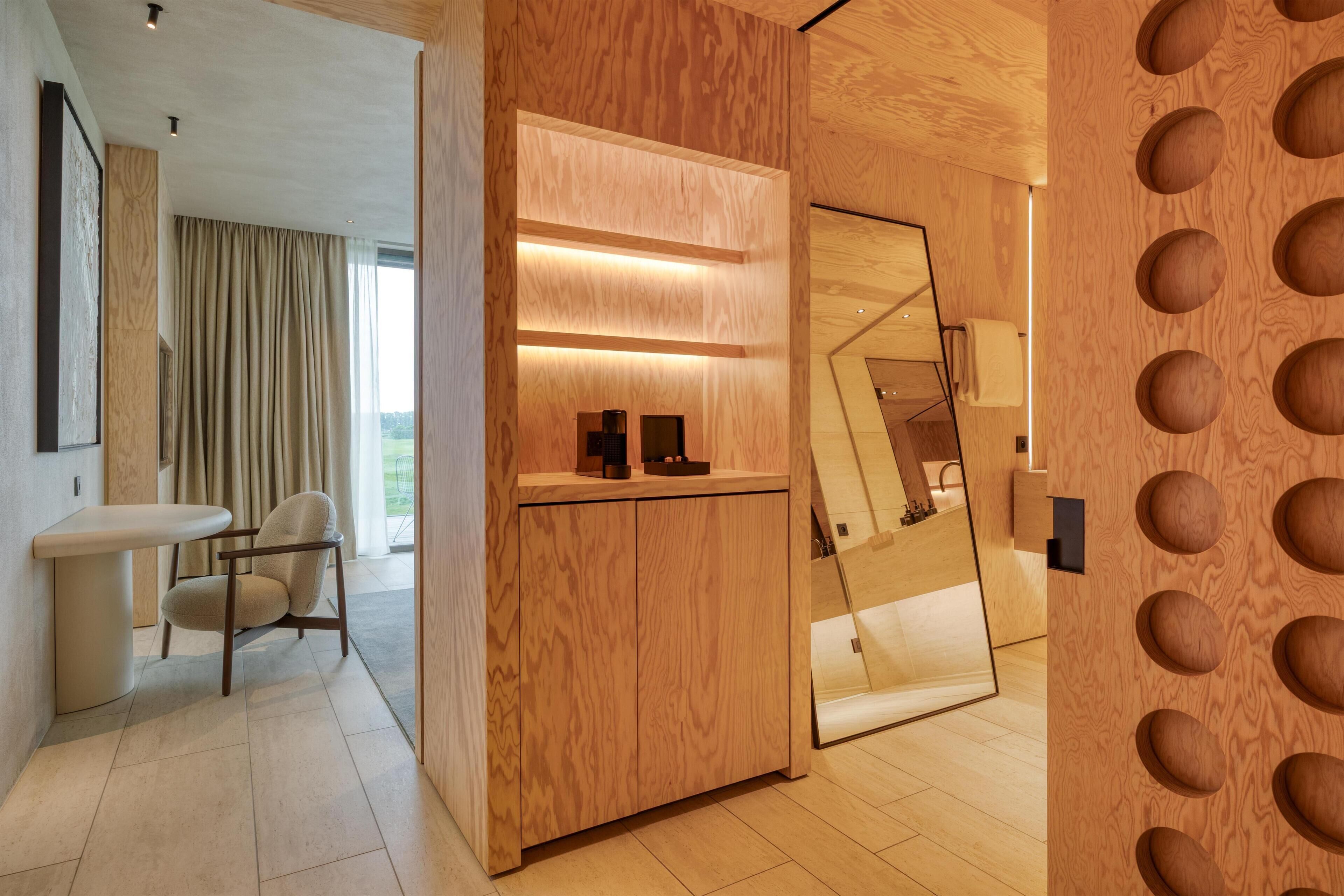 A modern hotel room with a wooden accent wall, a seating area with a chair, a small table, and a mirror.