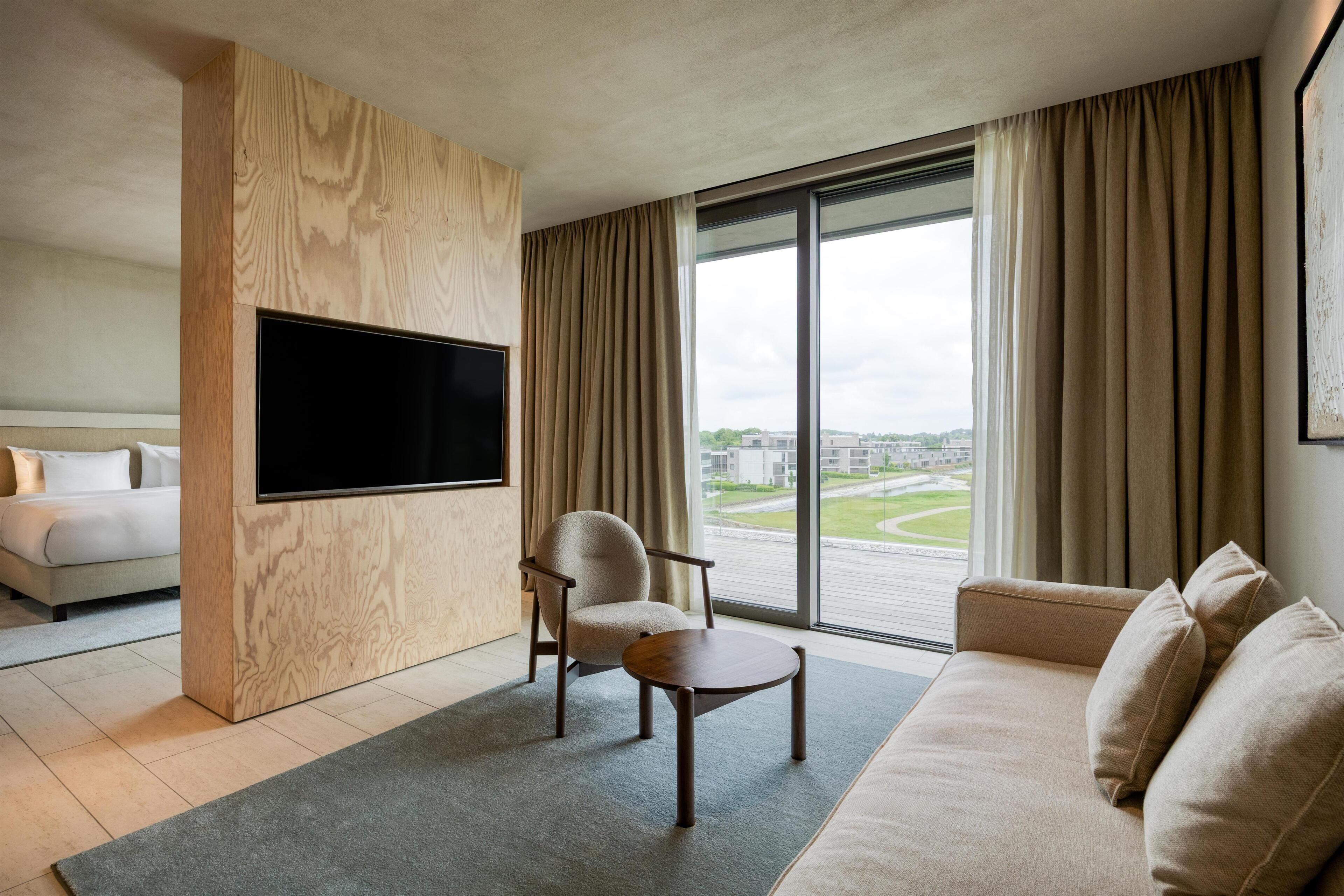 A modern hotel room with a large flat-screen TV, a sofa, a chair, and a view of the city through large windows.