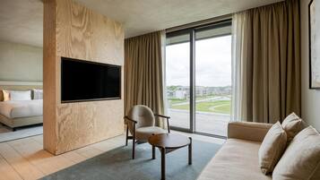 A modern hotel room with a large flat-screen TV, a sofa, a chair, and a view of the city through large windows.