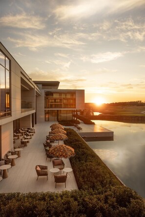 Un edificio moderno con una terrazza sul tetto, arredi esterni e ombrelloni, con vista su una superficie d'acqua tranquilla e un tramonto.