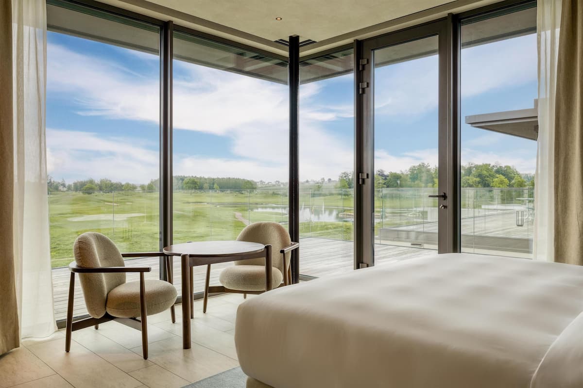 Ett modernt hotellrum med en stor säng, ett matbord, två stolar och utsikt över en golfbana genom stora fönster från golv till tak.