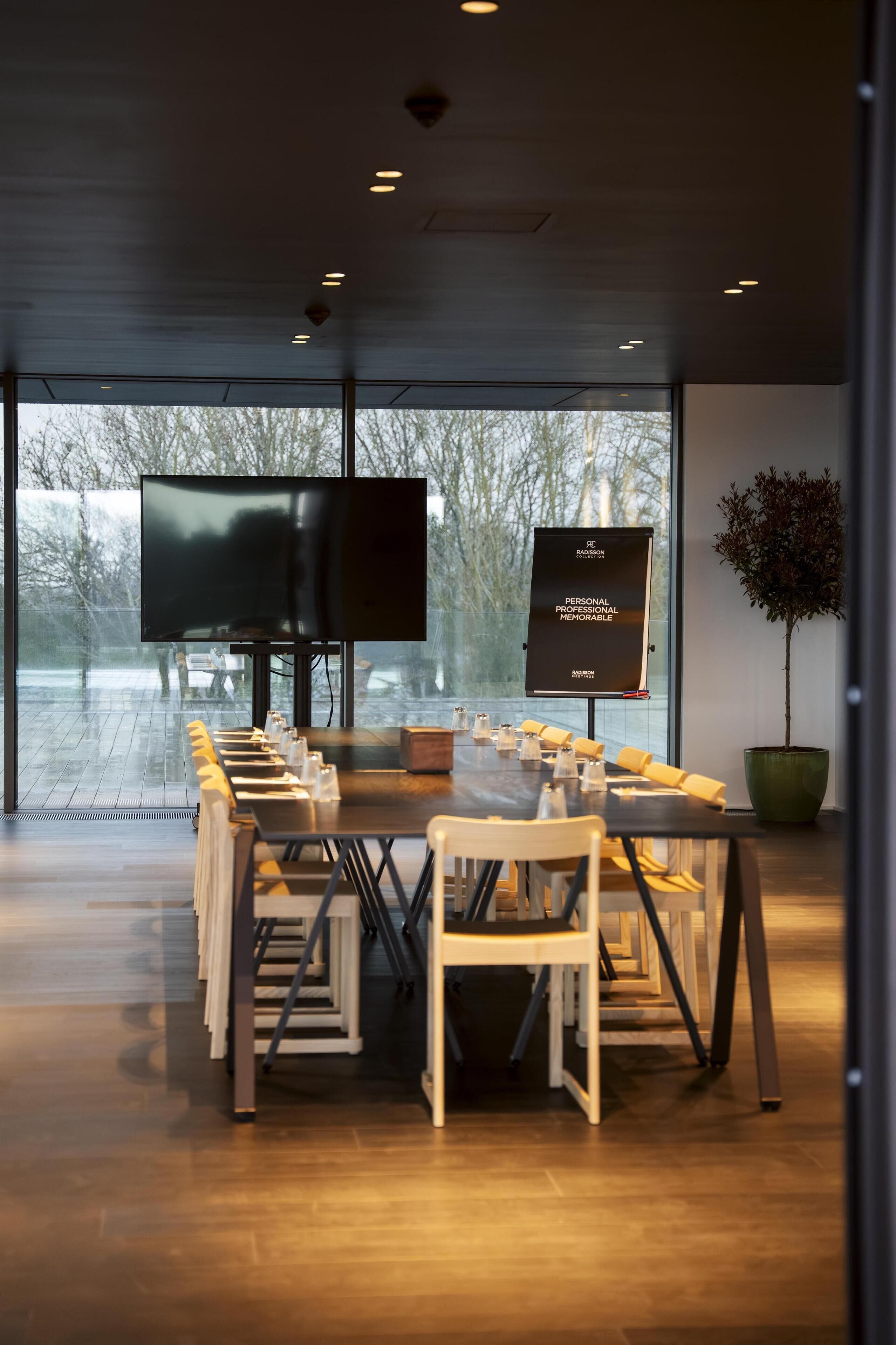 A conference room with a long table, chairs, a large screen, and a potted plant.