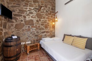 Une pièce avec un mur en pierre, un lit avec une literie blanche et des coussins à motifs, une barrique en bois, une petite table et un téléviseur fixé au mur.