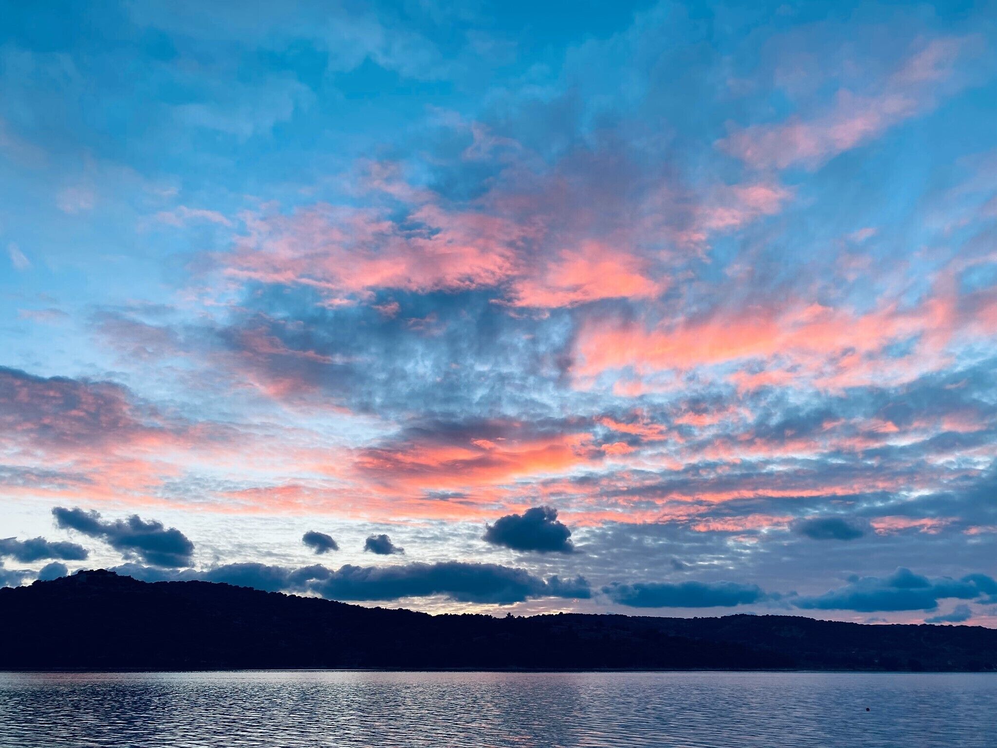 En rolig sø med silhuetten af et bjerglandskab i det fjerne og en himmel med spredte skyer.