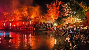 Evento en la playa con gente, un escenario y una masa de agua que refleja luces.