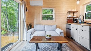 L’intérieur chaleureux d’un chalet avec un canapé, une table en bois sur laquelle se trouve un saladier, un coin cuisine et une fenêtre donnant sur les arbres.