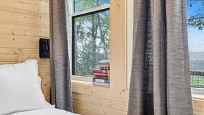 Une pièce rustique en bois, avec un lit, une fenêtre ornée de rideaux et une pile de livres posée sur le rebord de la fenêtre.