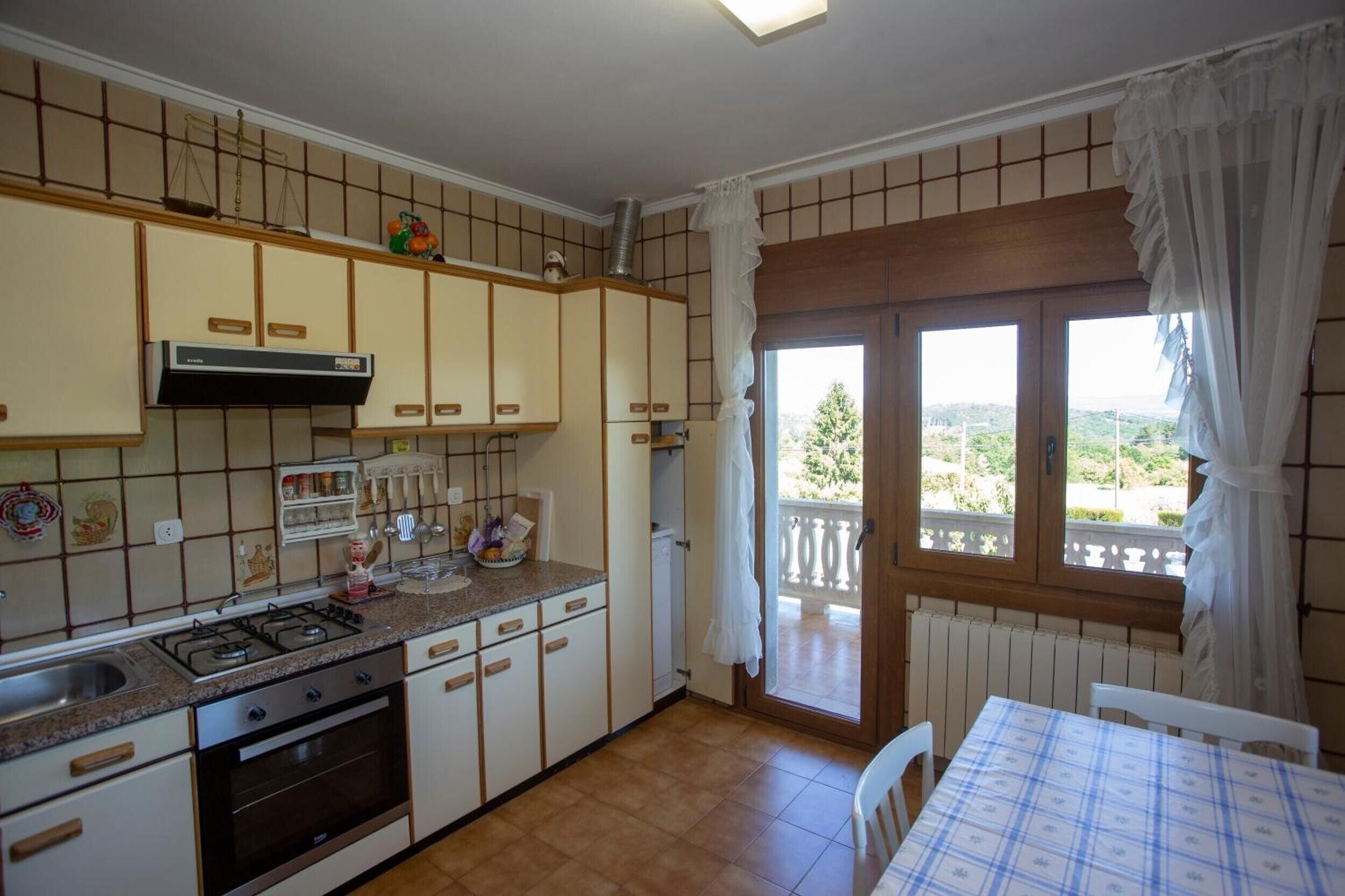 Uma cozinha com fogão a gás, forno e armários, uma mesa de jantar com toalha quadriculada e vista pela janela.