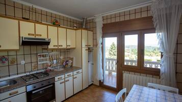 Uma cozinha com fogão a gás, forno e armários, uma mesa de jantar com toalha quadriculada e vista pela janela.