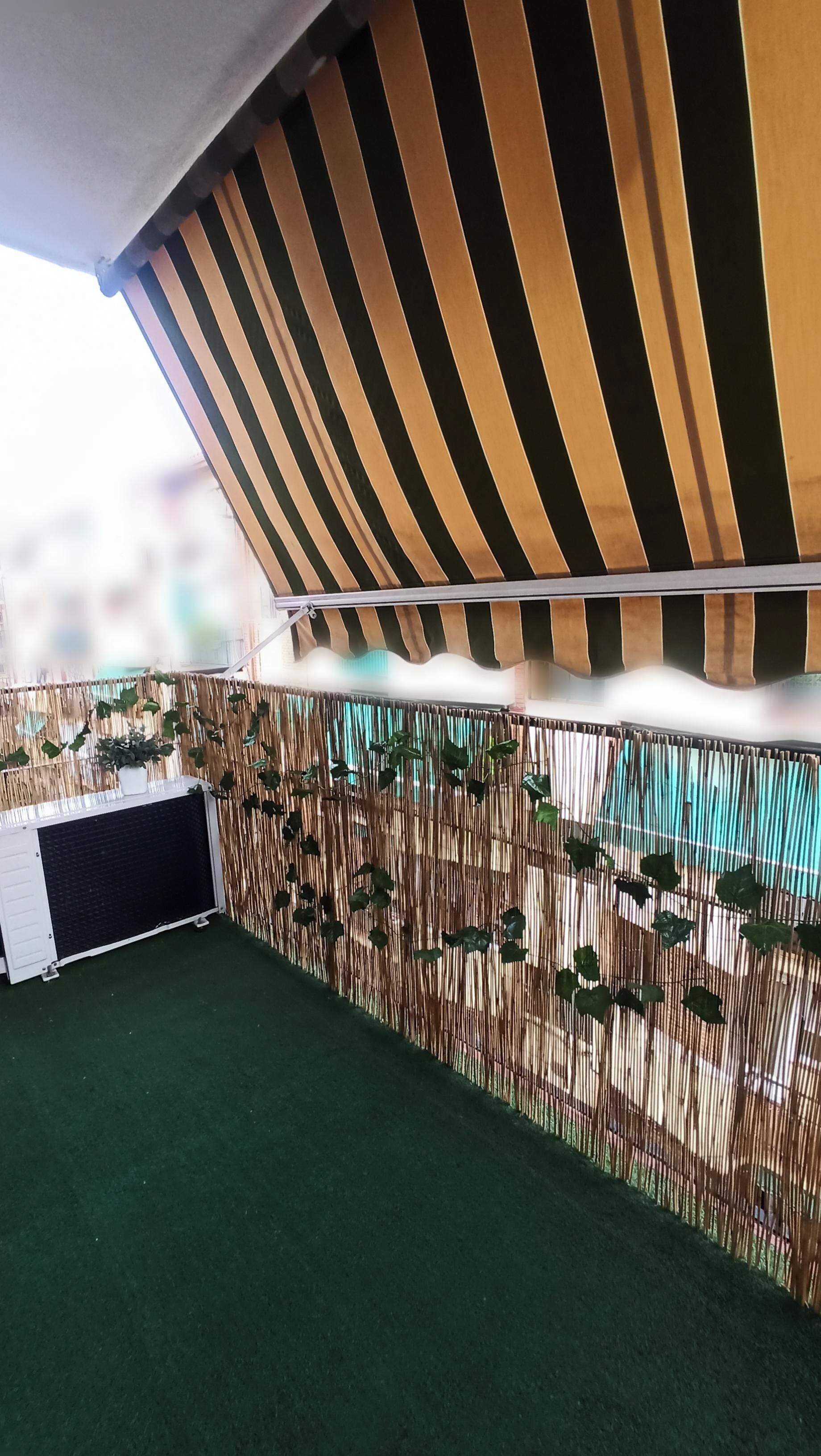 Terraza al aire libre con toldo a rayas, césped artificial y cerca de bambú con plantas colgantes.