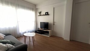 Ein modernes Wohnzimmer mit einer Couch, einem kleinen weißen Tisch, einem Fernseher auf einem weißen Lowboard und einer Tür.