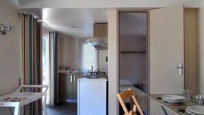 Una cocina compacta con refrigerador, estufa y alacenas. Se ve una mesa de comedor con sillas, y una cortina a rayas cuelga al fondo.