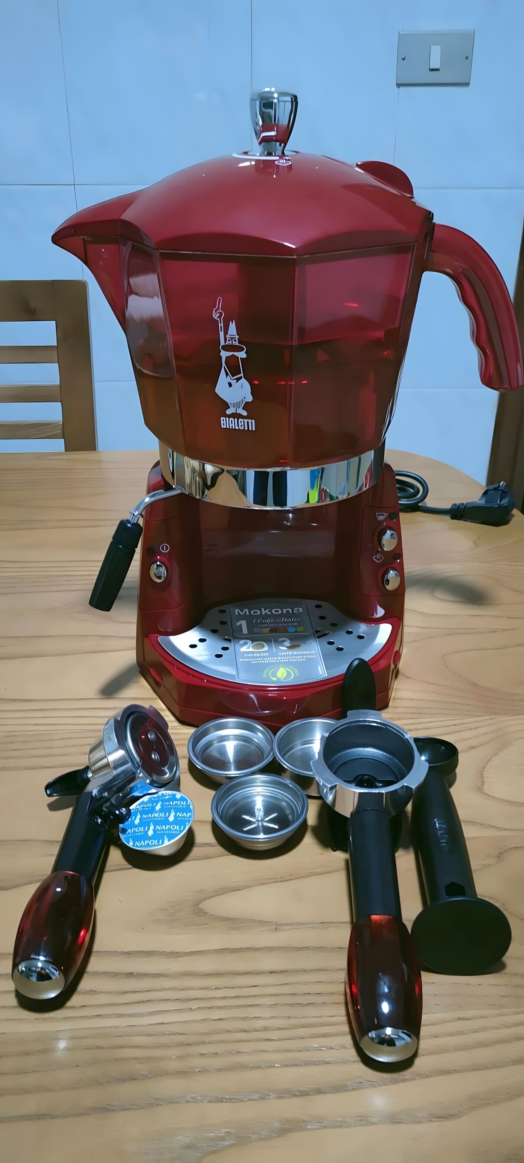 A red coffee machine with a logo, a control panel, and several coffee capsules on a wooden table.