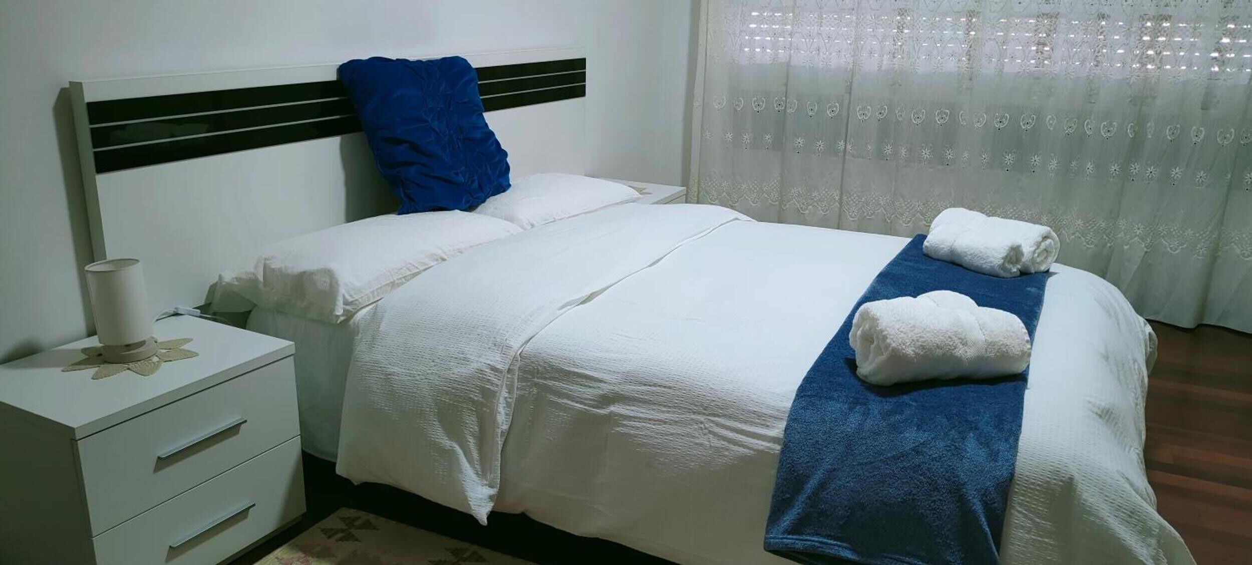 Un lit bien fait, avec du linge de lit blanc et une serviette bleue, une table de chevet avec une lampe, et une fenêtre ornée de rideaux légers.