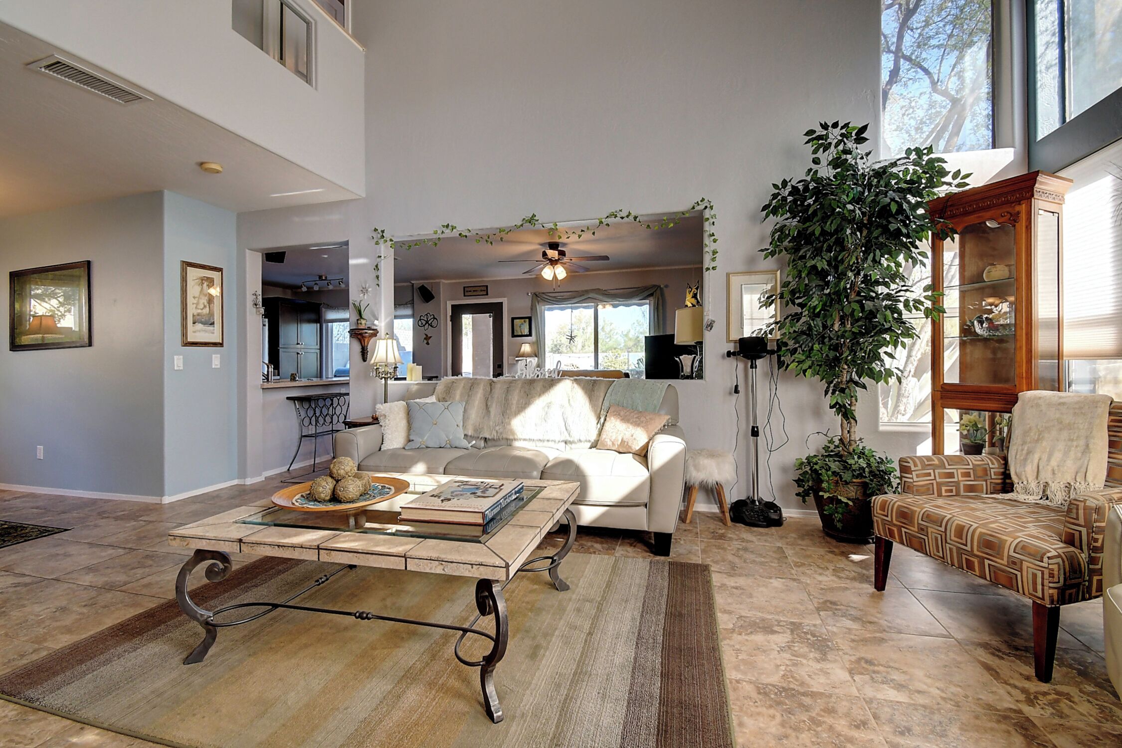 A spacious living room with a large sofa, a coffee table, and a patterned armchair. There is a large window providing natural light and a view of the outdoors.