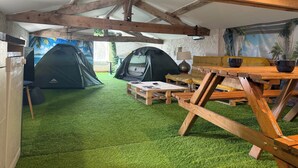 Un área de alojamiento con carpas, una mesa de madera y sillas, piso de césped artificial y un mural con paisaje tropical.