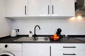 Une cuisine équipée d’un lave-linge, d’une machine à café et d’un panier de fruits posé sur le plan de travail.