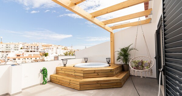 A rooftop terrace with a wooden hot tub, a hanging chair, and a view of a cityscape.