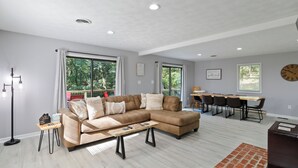 Amplia sala de estar con un sofá modular, un comedor con mesa y sillas, y ventanas grandes con vistas al exterior.