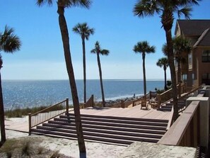 A beachfront property with a wooden staircase leading to a deck overlooking the ocean, surrounded by palm trees. - Oceanfront 1 Bedroom Condo - 644 Newhaven (Fripp Island)