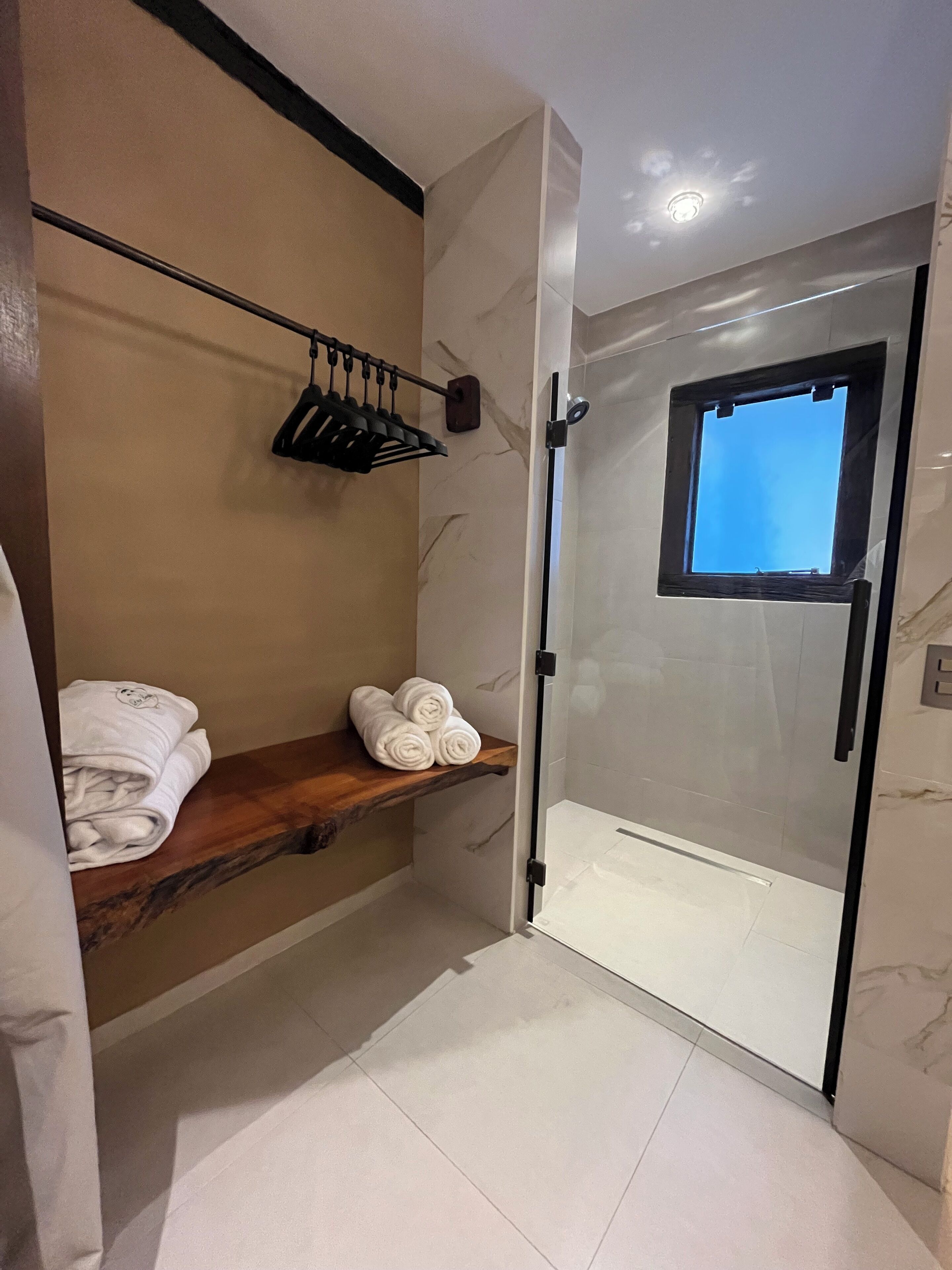 Un baño moderno con mampara de vidrio, repisa de madera con toallas y un ventanal pequeño.